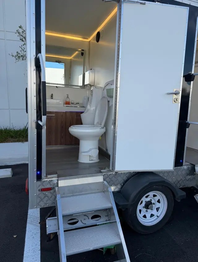 Restroom trailer door open showing toilet, vanity and metal steps